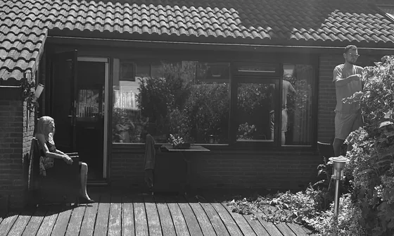 Trudy zit in de zon terwijl haar zoon en kleinzoon het tuinwerk doen.
